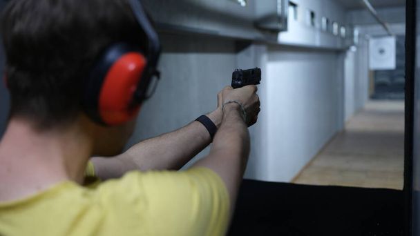 Крупный план мужчины-стрелка в наушниках, нацеленного на цель из пистолета в тире. Стрельба практикуется с оружием в стрельбище. Экстремальная концепция хобби - Фото, изображение