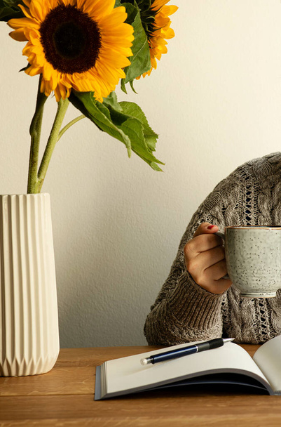 Woman with cup of coffee in her hand. Notebook, pen and vase with fresh sunflowers on the wooden table. Concept of inspirational workplace when working remote, studying or at home office. - Photo, Image