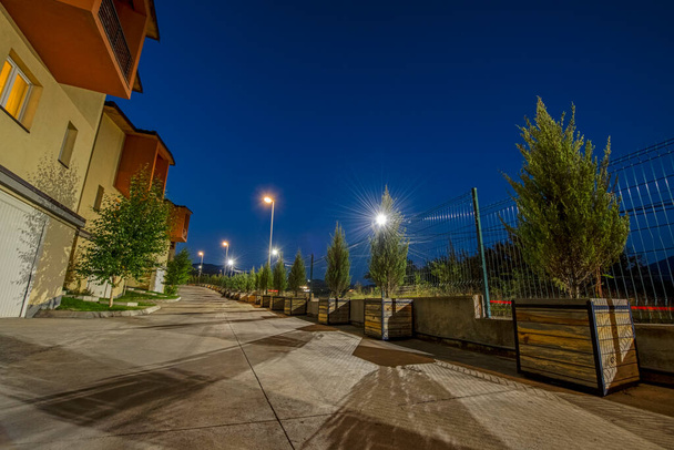 Wide angle night street scene with orange buildings, row of green conifer trees in wooden pots and red motion blur car lights under the navy blue sky with stars. Long exposure photography - Foto, Imagem