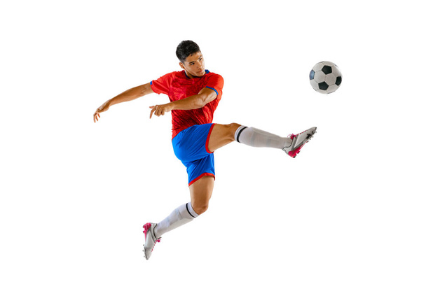 Portrait of young man in uniform, professional football player kicking ball in a jump isolated over white studio background. Concept of sport, team game, action, motion. Copy space for ad, poster - Foto, afbeelding