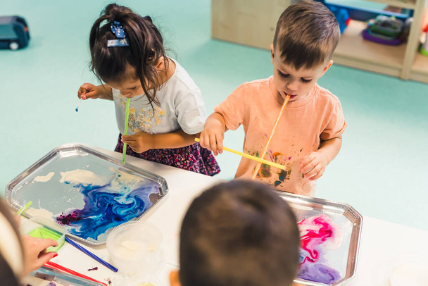 Многокультурная детская школа. Тоддлеры играют с полосатыми соломинками и рисованием молоком, используя нетоксичную пищевую окраску для цветов. Творческая активность детей для развития мозга детей. Мелкая моторика - Фото, изображение
