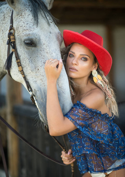 Beautiful blonde woman with curly hair, hat and horse. Portrait of a girl with white bikini ,blue transparent blouse  and her horse. Beautiful girl interacting and having fun with a horse at the ranch - Foto, immagini