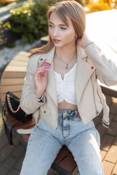 Fashionable young beautiful woman model with fashion streetwear with leather jacket and blue stylish jeans with handbag sits on the wooden bench near a cafe at sunset - Foto, imagen