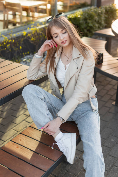 Stylish beautiful young woman hipster in fashionable rock streetwear with leather jacket, top, blue vintage jeans and white sneakers sits on wooden bench near a cafe - Foto, afbeelding