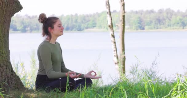 Gros plan d'une jeune femme pratiquant le yoga dans la nature. Asseyez-vous sur l'herbe verte à côté d'un arbre et d'un lac forestier. Communiquer pour faire de l'exercice et maintenir une bonne santé. Images 4k de haute qualité - Séquence, vidéo