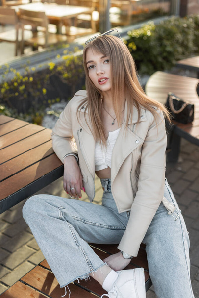 Glamorous pretty woman hipster model with fashion casual rock style with leather jacket and vintage blue jeans sits on bench on the street - Foto, Bild