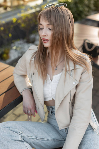 Trendy beautiful young model girl in fashionable casual autumn outfit with a rock leather jacket, white top and jeans sits on a bench outside a cafe on the street - Photo, image