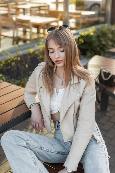 Fashionable beautiful female portrait of a pretty young hipster girl in a street fashion outfit with leather rock jacket and blue jeans sitting on a bench outside a cafe in the city - 写真・画像