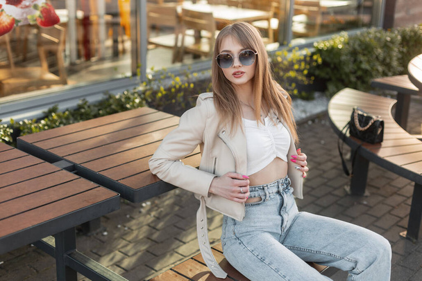 Fashionable beautiful stylish hipster girl model with round  sunglasses in a fashion leather rock jacket with a top and vintage blue jeans and a bag sitting on a wooden bench near a cafe at sunset - Foto, imagen