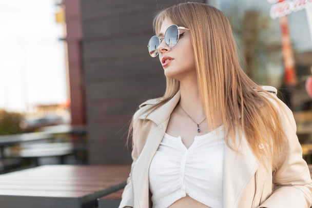 Уличный портрет модной красивой молодой женщины-хипстера со стильными круглыми солнцезащитными очками в модной скальной коже и белой майке сидит рядом с кафе в городе - Фото, изображение