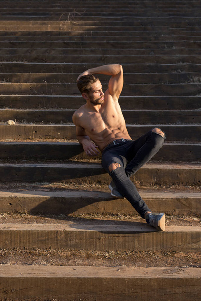 Full body of serious young muscular male with naked torso in stylish ripped jeans touching head and looking away while sitting on stairway at sunset in park - Foto, Imagem