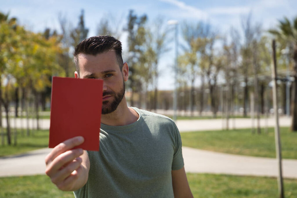 Young and handsome man, with a beard and green shirt, showing a red card with a serious gesture. Concept sport, expulsion, rejection, football, world, competition. Selective focus. - Foto, immagini