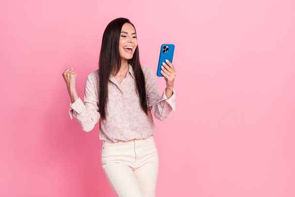 Photo portrait of excited young lady hold gadget raise fist job promotion wear stylish formalwear look isolated on pink color background. - 写真・画像