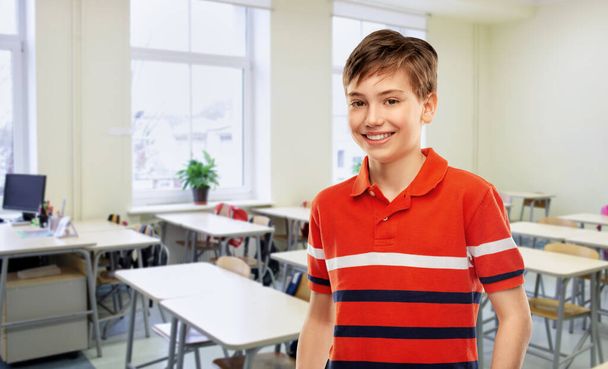 school, education and people concept - portrait of happy smiling student boy in red polo t-shirt over empty classroom background - Foto, Imagem