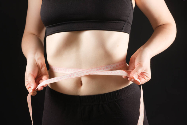 Eine Frau misst den Bauchumfang auf schwarzem Grund. Das Konzept der Gewichtsabnahme. - Foto, Bild