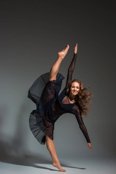Молодая красивая танцовщица в черном платье позирует на темно-сером фоне студии. Современная, современная, импровизация - Фото, изображение