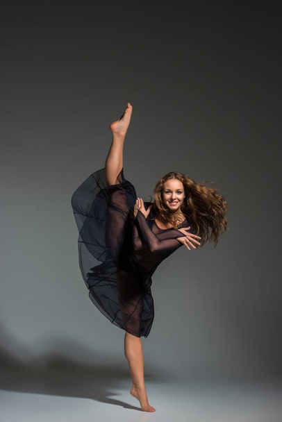 Νεαρή όμορφη χορεύτρια με μαύρο φόρεμα ποζάρει σε σκούρο γκρι φόντο στούντιο. Σύγχρονη, Σύγχρονη, αυτοσχεδιασμός - Φωτογραφία, εικόνα