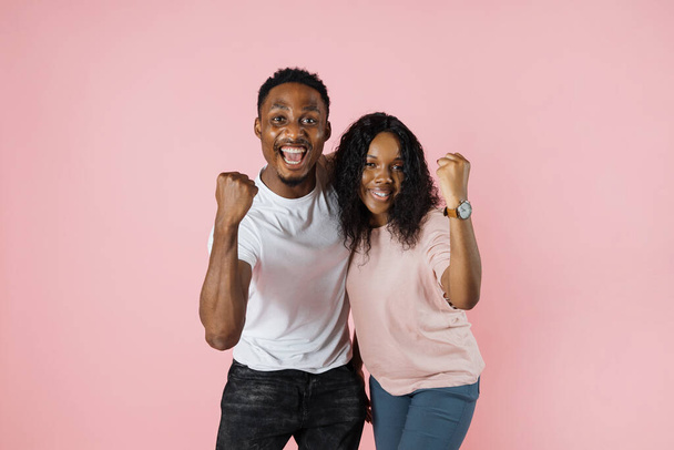 Big win. Happy excited african couple celebrating success with raised fists. Cheerful african american man and woman exclaiming with joy while standing isolated over pink background, copy space. - Fotó, kép