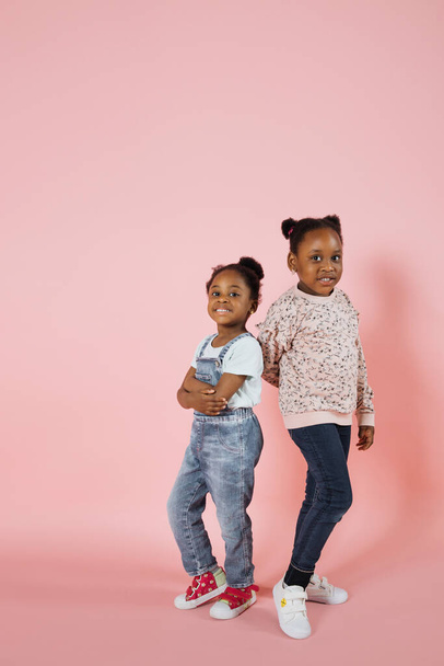 Happy childhood, family concept. Full length portrait of two cute adorable African kids girls, standing back to back having fun together. Isolated on pink background. - Valokuva, kuva