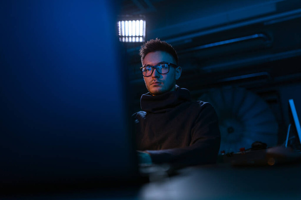 cybercrime, hacking and technology concept - young man in glasses writing code on laptop in dark room - Foto, Imagem