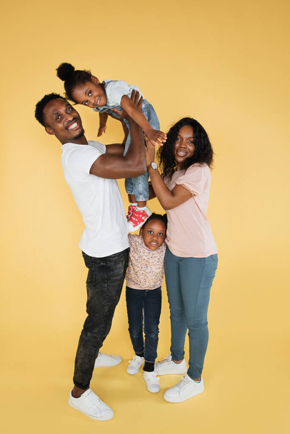 Cheerful african man holding excited daughter on hands. Woman and another daughter looking posing at camera, standing isolated over yellow studio background, panorama, banner, free copy space - 写真・画像
