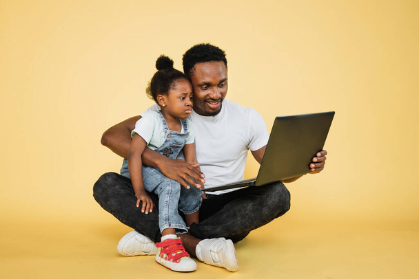 Horizontal shot of happy joyful African family, young father with cute little upset kid daughter, sitting on the floor on yellow studio background and watching a video lesson on laptop pc. - Foto, Bild