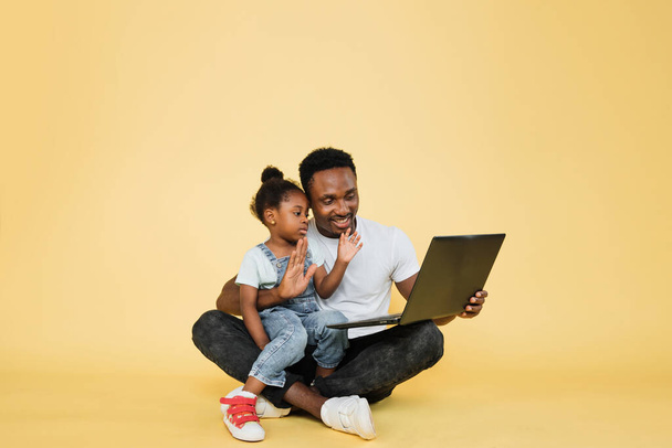 Horizontal shot of happy joyful African family, young father with cute little kid daughter, sitting on the floor and waving hands while watching a video lesson on laptop pc on yellow studio background - Valokuva, kuva