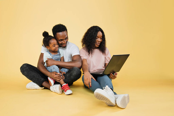 Happy family, technology, communication concept. Young african parents sitting on floor with their daughter and embracing, using laptop for video call, over yellow studio background, free space. - Foto, afbeelding