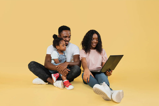 Happy family, technology, communication concept. Young african parents sitting on floor with their daughter and embracing, using laptop for video call, over yellow studio background, free space. - Foto, imagen