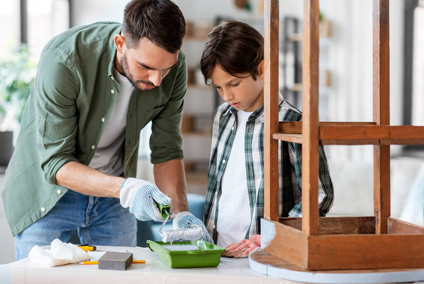 renovation, diy and home improvement concept - father and son in gloves with paint roller painting old wooden table in grey color - 写真・画像