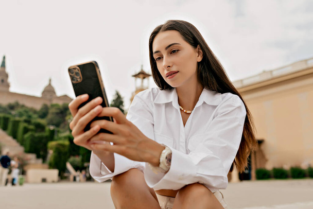 Successful elegant european woman with dark loose hair wearing white shirt using smartphone while sitting on stairs on background of city - Fotoğraf, Görsel