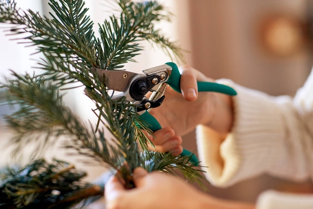 winter holidays, diy and hobby concept - close up of woman with fir branches and pruner making christmas wreath at home - Fotó, kép