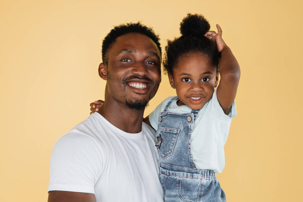 Family, fathers day concept. Close up studio portrait of happy young dark skinned daddy with his cute little daughter, smiling, hugging and having fun together, on isolated yellow background. - Foto, Bild