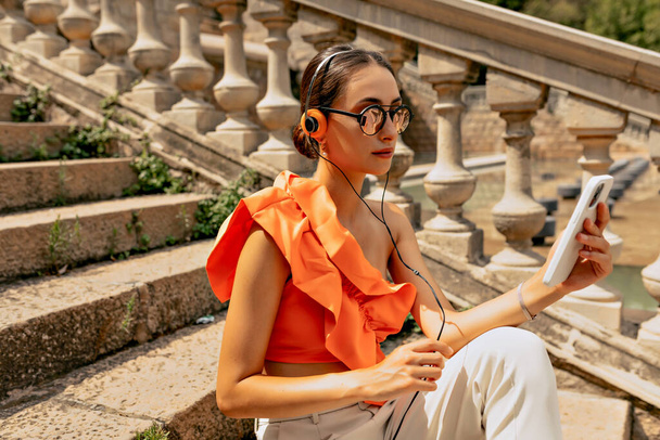 An elegant young brunette in cool glasses sits on the stairs in headphones and looks into a smartphone. Engaging young dark-haired girl in bright orange blouse, modern sunglasses posing outdoors. - Foto, Bild