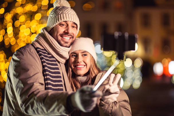 winter holidays and people concept - happy smiling couple taking picture with smartphone on selfie stick and hugging over christmas lights in evening - Φωτογραφία, εικόνα