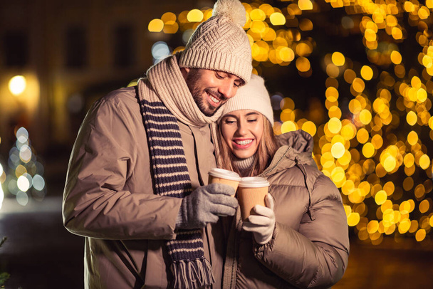 winter holidays, hot drinks and people concept - happy young couple with takeaway coffee cups in christmas city in evening - 写真・画像