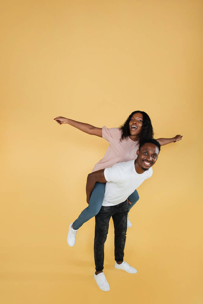 I will show you sky. Portrait of positive cheerful african american man hold his girlfriend piggyback imagine fly like airplane wear casual style clothes isolated over yellow color background. - Foto, afbeelding