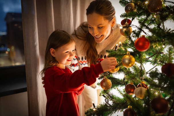 family, winter holidays and people concept - happy mother and little daughter decorating christmas tree at home - Zdjęcie, obraz
