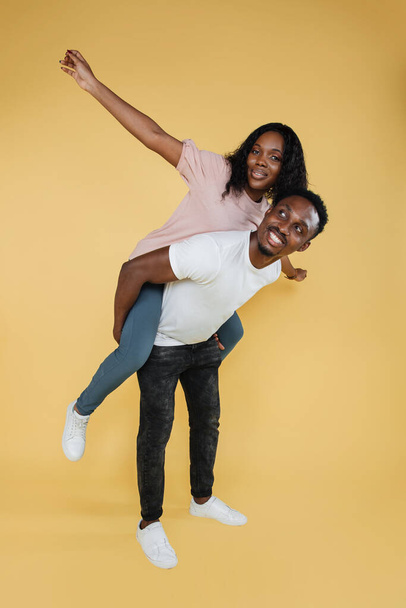 I will show you sky. Portrait of positive cheerful african american man hold his girlfriend piggyback imagine fly like airplane wear casual style clothes isolated over yellow color background. - Foto, Imagen