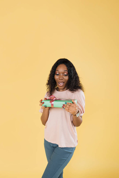 Portrait of excited funny african american woman with afro hair, wearing pink t-shirt, posing with gift box holding in her hand, isolated on yellow background. - Foto, imagen