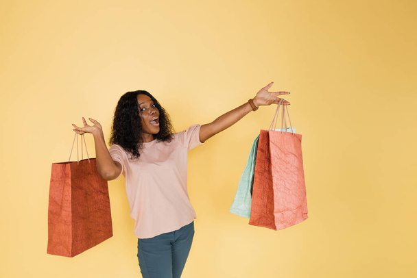 Portrait of positive cheerful afro american girl, addicted shopper, demonstrating a lot of colorful shopping bags to camera, while standing on yellow background, copy space. - Foto, afbeelding