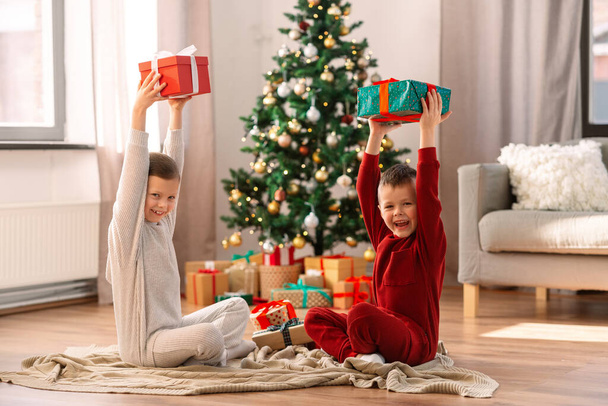 christmas, winter holidays and childhood concept - happy girl and boy in pajamas with gifts sitting on floor at home - Fotó, kép