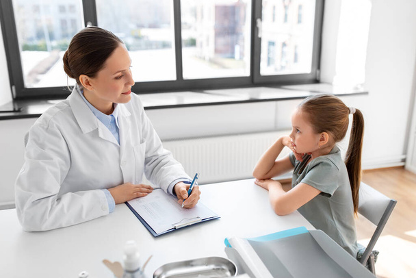 medicine, healthcare, pediatry and people concept - female doctor or pediatrician with clipboard talking to little girl patient on medical exam at clinic - Foto, immagini