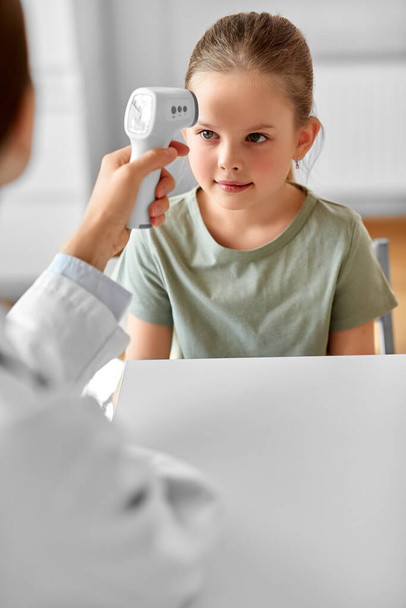 medicine, healthcare and pediatry concept - female doctor or pediatrician measuring little girl patients temperature with infrared forehead thermometer at clinic - Foto, imagen