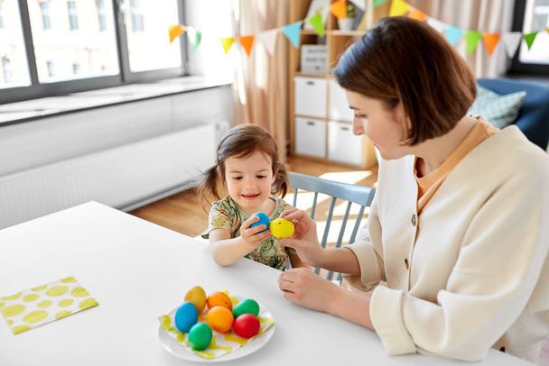 vacaciones, la familia y el concepto de la gente - la pequeña hija feliz bebé y la madre tocando huevos de Pascua de colores en casa - Foto, imagen