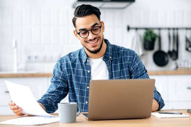 Work from home. Smart arabian or indian stylish man, freelancer, programmer, manager, working remotely, sit at a desk at home in the kitchen, working on a project uses laptop, studying documents,smile - Foto, imagen