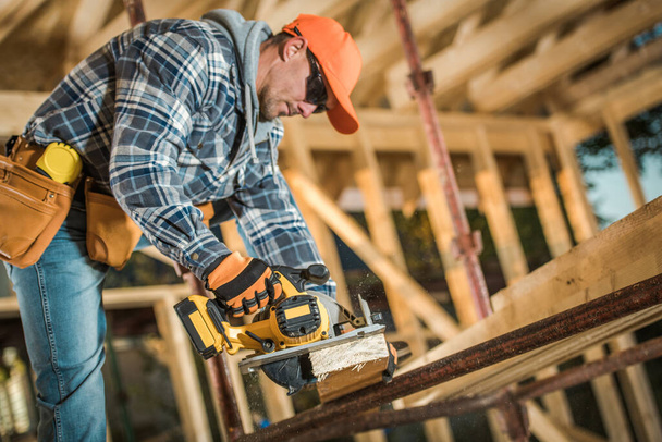 Caucasian Middle Aged Builder Working with Electric Wood Planer Power Tool at the Construction Site of Wooden Skeleton of Canadian Style Residential Building. Industrial Theme. - 写真・画像