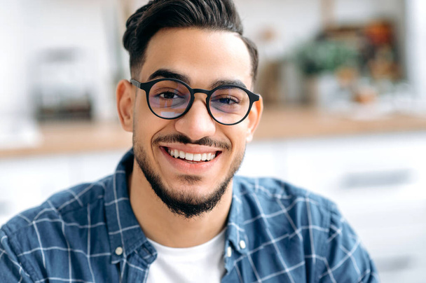 Close-up photo of a handsome attractive mixed race young man with glasses, freelancer or student, in casual shirt, with perfect white teeth, looking at the camera with toothy happy smile - Foto, afbeelding