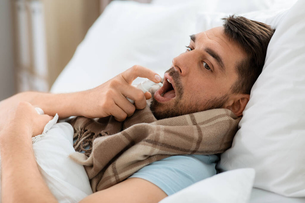 people and health problem concept - unhappy sick man spraying his throat with oral spray lying in bed at home - Foto, imagen