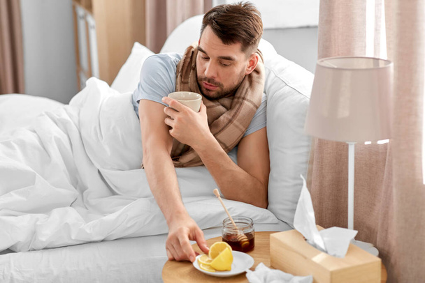 health, illness and people concept - sick man drinking hot tea with lemon and honey in bed at home - Photo, Image
